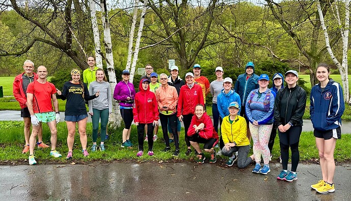 A group of runners in bright clothing stand next to trees and a paved walking path.