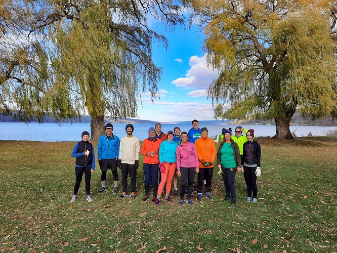 A group of runners wearing bright colored clothes stand in front of two trees and a lake.